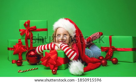 little girl in a Santa hat is lying on a gift box with a red bow on a green background in the Studio. the child smiles happily and looks up. Advertizing. Copy space.
