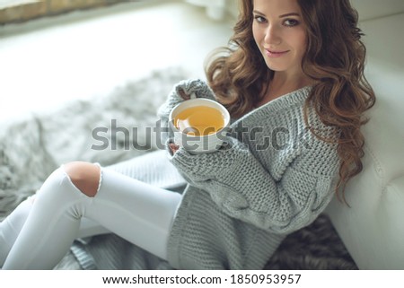 Close-up of female hands with a mug of drink. Beautiful girl in a gray sweater is holding a cup of tea or coffee in the morning sunlight. Mug for your design. High quality photo.