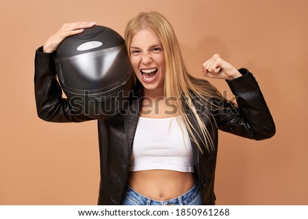 Ecstatic young blonde woman biker in stylish leather jacket keeping fist clenched and mouth wide opened, screaming, expressing true emotions, holding protective helmet. Success, triumph and winning
