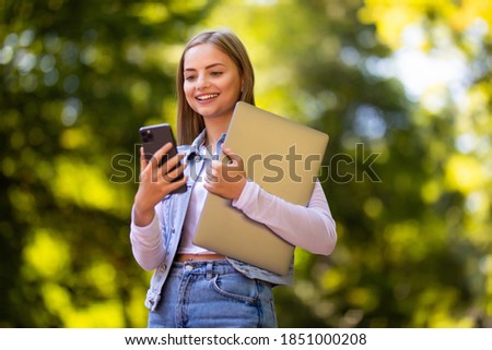 Photo of happy woman walking in park outdoors holding laptop computer looking aside chatting by mobile phone.