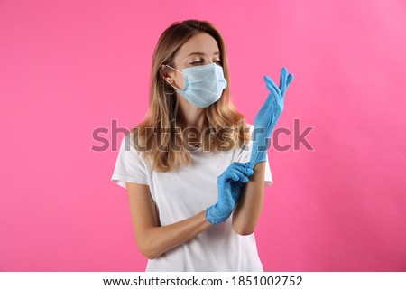 Young woman in protective mask putting on medical gloves against pink background