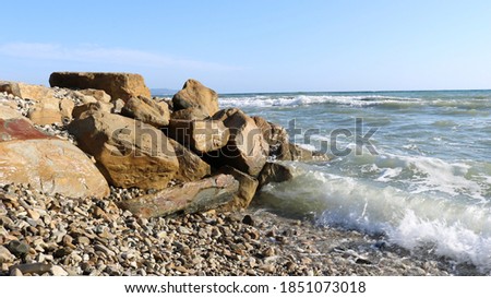 greenish waves rolling on a rocky shore with large brown stones against a bright blue sky, the image of a sunny sea and a deserted beach on a clear day
