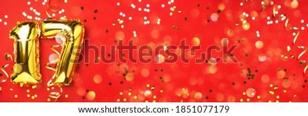 Gold foil balloon number, digit seventeen. Birthday greeting card, inscription 17. Anniversary celebration event. Banner. Stylish golden numeral, light bokeh, glitter, red background. Numerical digit.