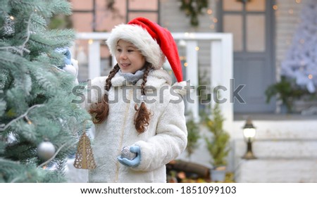 Happy girl in a white fur coat and with a santa hat outdoors decorates the Christmas tree against the background of the Christmas house. Happy New Year and Merry Christmas!