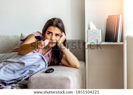 Photo of a young woman sitting on a sofa, feeling depressed and changing channels on a TV remote. Young brunette woman relaxing at home, curled up in a blanket, holding the remote, watching television