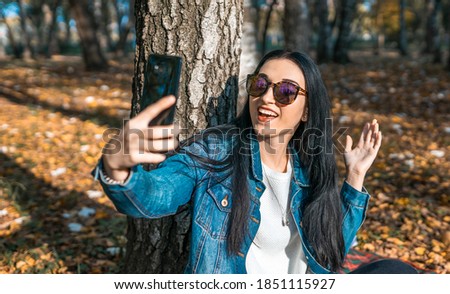 Attractive young woman taking selfie on a mobile phone in a yellow autumn park. A cute woman takes pictures of herself while walking outdoors on a sunny autumn day. Blogging concept