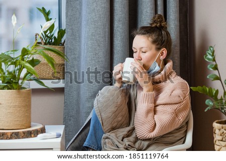 Sick European attractive young woman has a cold and drinks hot tea upset young blonde in a sweater and blanket feels bad, puts on a medical mask. cold, nausea, chills.