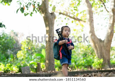 An Asian toddler girl is carrying a camera tripod. she is wearing a navy blue shirt and carrying a green-pink bag by herself. she is looking for something.