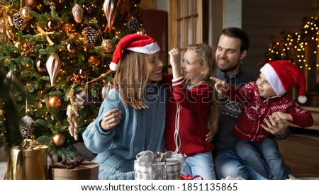 Wide banner view image of happy young Caucasian family with two small kids celebrate New Year at home. Smiling parents have fun sit on floor near Christmas tree, enjoy winter holidays with children.