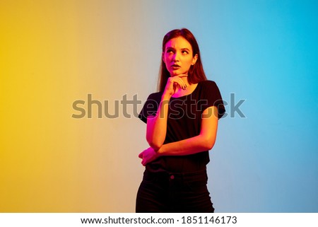 Thoughtful, dreamful. Young caucasian woman's portrait on gradient blue-yellow studio background in neon light. Concept of youth, human emotions, facial expression, sales, ad. Beautiful brunette model