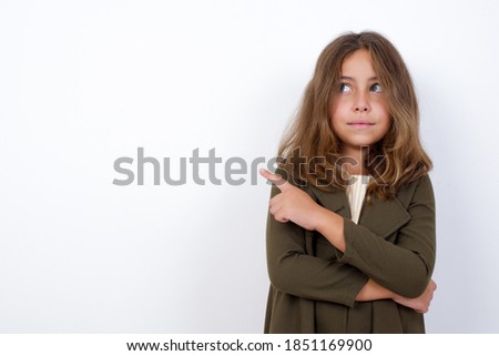 Portrait of Young beautiful Caucasian little girl standing against white background posing on camera with tricky look, presenting product with index finger. Advertisement concept.