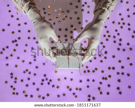 Hands in rubber protective gloves hold a white gift box. Golden and silver confetti stars glitter on a pink background. Holiday concept in the context of the coronavirus pandemic.