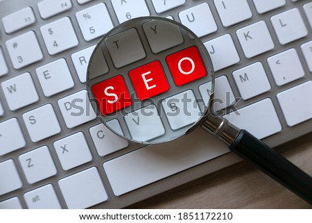 Selective focus of magnifying glass and computer keyboard with red key written with SEO Search Engine Optimization on a wooden background. Royalty-Free Stock Photo #1851172210