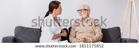 geriatric nurse talking to aged man while sitting on sofa at home, banner