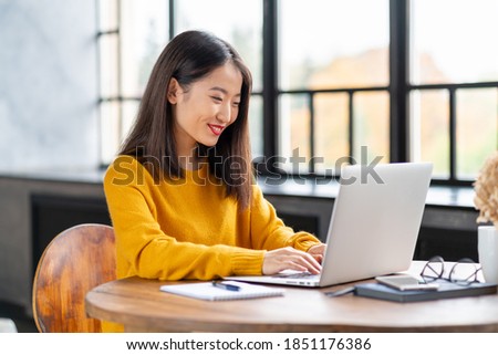 Asian woman working on laptop at home or in cafe. Young lady in bright yellow jumper sitting at desk typing on computer. Business oriental female in front of window