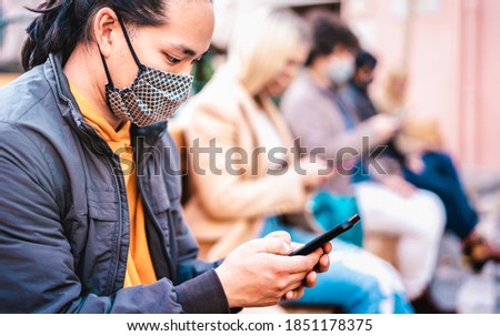 Asian guy using smart phone covered by face mask on Covid second wave - New normal lifestyle concept with milenial people watching news on mobile smartphone - Shallow depth of field with focus on man
