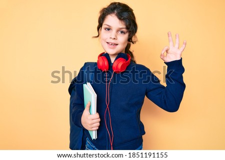 Cute hispanic child holding student backpack and books doing ok sign with fingers, smiling friendly gesturing excellent symbol 