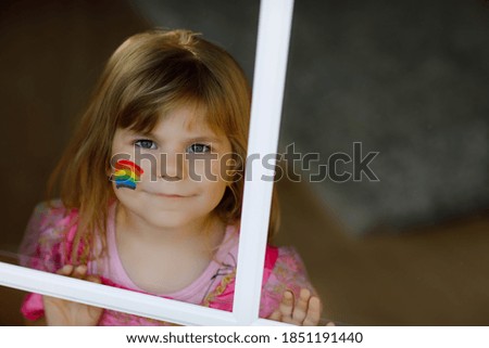 Cute lonely little toddler girl in princess dress sitting by window with rainbow with colorful colors on face during pandemic coronavirus quarantine. Children make and paint rainbows around the world