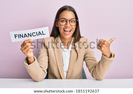Beautiful hispanic business woman holding financial message paper pointing thumb up to the side smiling happy with open mouth 