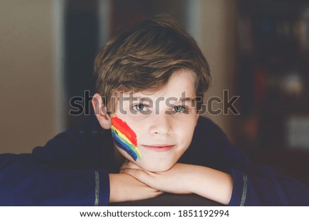 Portrait of adorable school kid boy with painted rainbow with colorful colors on face. Lonely child during pandemic coronavirus quarantine. Children make and paint rainbows around the world