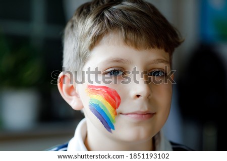 Portrait of adorable school kid boy with painted rainbow with colorful colors on face. Lonely child during pandemic coronavirus quarantine. Children make and paint rainbows around the world