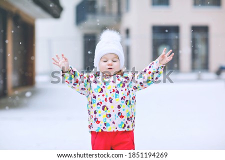 Portrait of little toddler girl walking outdoors in winter. Cute toddler eating sweet lollypop candy. Child having fun on cold snow day. Wearing warm baby colorful clothes and hat with bobbles.