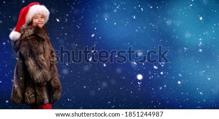 Christmas winter kids fashion. Cute little girl in a silver fox fur coat and a Santa Claus hat stands under the snowfall on a dark blue background and laughs. Copy space. 
