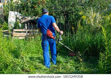 Funny fat man in a blue jumpsuit and baseball cap, mowing the grass with a trimmer.