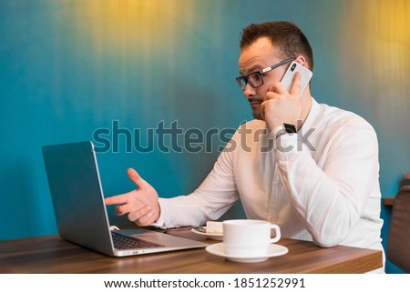 Portrait of a young attractive businessman in a white shirt, glasses, talking on the phone and looking at a laptop with a cup of coffee in a cafe