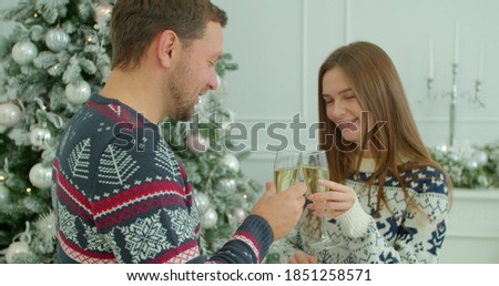 Couple near glowing fir tree holding champagne glasses saying a toast best wishes celebrate Christmas together. Romantic date on xmas eve at modern decorated house.