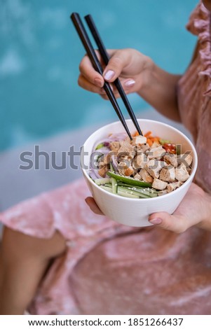 Woman sitting by the pool against blue water background with chopsticks and eating takeaway healthy organic Hawaiian chicken poke bowl with cucumber, rice, carrots, onion. Healthy lifestyle concept