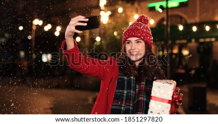 Close up portrait of young Caucasian female in red taking selfie photos on smartphone while standing in decorated with xmas lights city and smiling. Joyful Caucasian woman taking pictures on cellphone