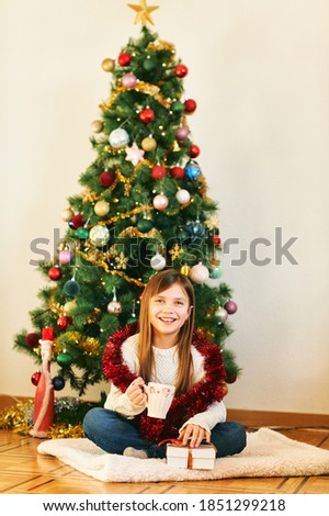 Portrait of sweet little girl sitting next to Christmas tree, wearing white cozy pullover, holding cup with hot chocolate, winter holidays