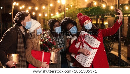 Portrait of happy mixed-races people in medical masks standing outdoor with x-mas presents and taking selfie photos on smartphone while snowing Caucasian and African American friends in decorated city