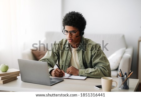Industrious black teenager in headset studying remotely from home, writing in notebook. African American student taking online class on laptop, watching video lesson, participating in conference Royalty-Free Stock Photo #1851316441