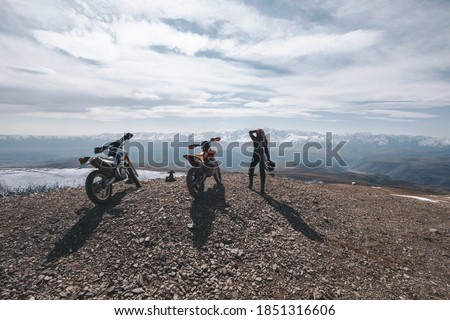 Female motorcyclist standing with her enduro motorcycle on mountain top, snow peaks skyline view