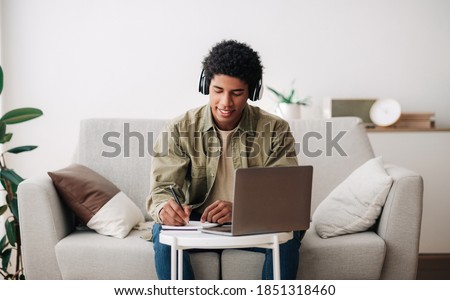Web-based education concept. Positive teen student in headphones writing in notebook during online lecture on laptop. Cool African American guy doing homework and listening to webinar, panorama Royalty-Free Stock Photo #1851318460