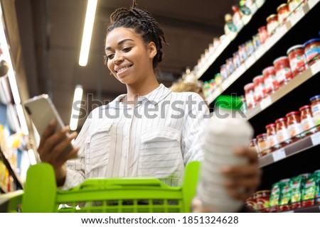 Happy Black Lady Using Cellphone With Grocery Shopping Checklist Application Buying Food Products In Supermarket Groceries Shop, Standing Wih Trolley Cart In Store. Selective Focus Royalty-Free Stock Photo #1851324628