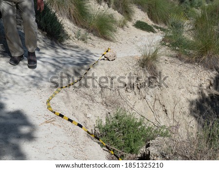 A hiker walks in shadow as he passes close a hazard point where the path is narrow and drops away steeply. The dangerous section is marked temporarily by simple yellow and black warning tape
