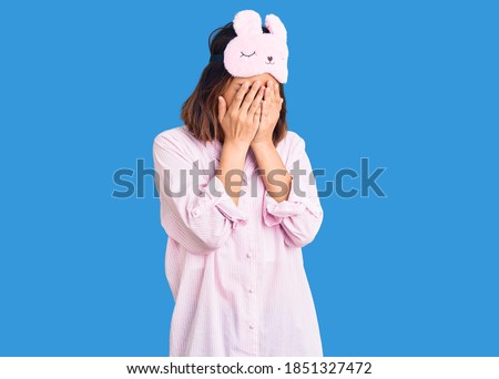 Young beautiful chinese girl wearing sleep mask and pajama with sad expression covering face with hands while crying. depression concept. 