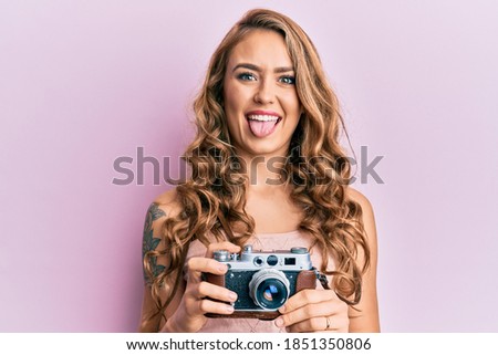 Young blonde girl holding vintage camera sticking tongue out happy with funny expression. 