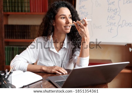 Technology And Communication Concept. Portrait of young woman sitting at desk, talking on mobile phone, using laptop and virtual voice assistant for online search or to record reminder