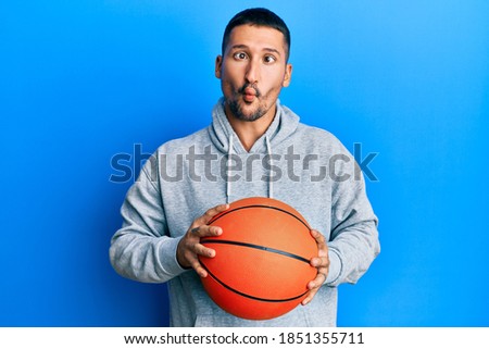 Handsome man with tattoos holding basketball ball making fish face with mouth and squinting eyes, crazy and comical. 