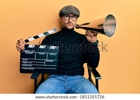 Handsome man with tattoos holding video film clapboard and louder skeptic and nervous, frowning upset because of problem. negative person.  Royalty-Free Stock Photo #1851355726