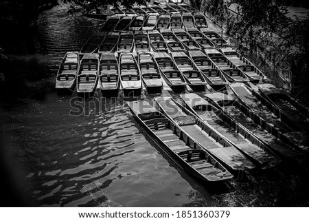 Black and white photo of punting boats by Magdalen Bridge Boathouse on river Cherwell in Oxford, many boats docked together in rows.