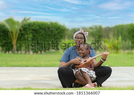 A black American daughter sitting on her mother's lap Playing an acoustic guitar in the park.