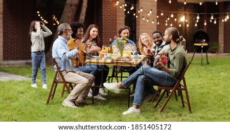 Mixed-races happy big family sitting at table with dinner at back yard outdoors and having fun. Guy playing on guitar. Musician entertaining at picnic. Royalty-Free Stock Photo #1851405172
