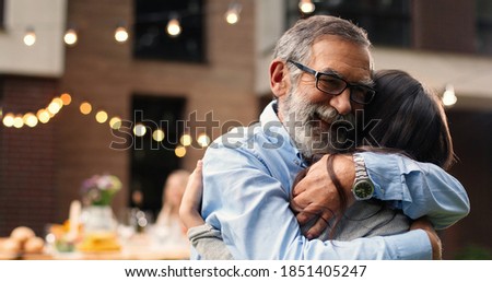 Close up of Caucasian happy small cute granddaughter hugging her grandfather in yard. Barbecue meal on background. Little girl embracing grandpa in glasses outdoors. Meeting concept. Royalty-Free Stock Photo #1851405247