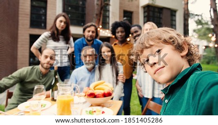 POV of small boy taking selfie photo with multi ethnic family smiling and waving to camera while having lunch at back yard at table. Picture of mixed-races people and kid.
