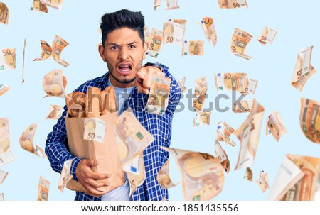 Handsome latin american young man holding paper bag with bread pointing displeased and frustrated to the camera, angry and furious with you
